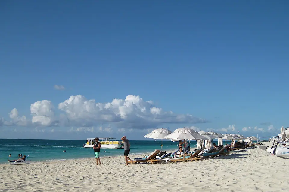 Guests walking past white umbrellas and loungers on Grace Bay Beach with turquoise water and a boat offshore