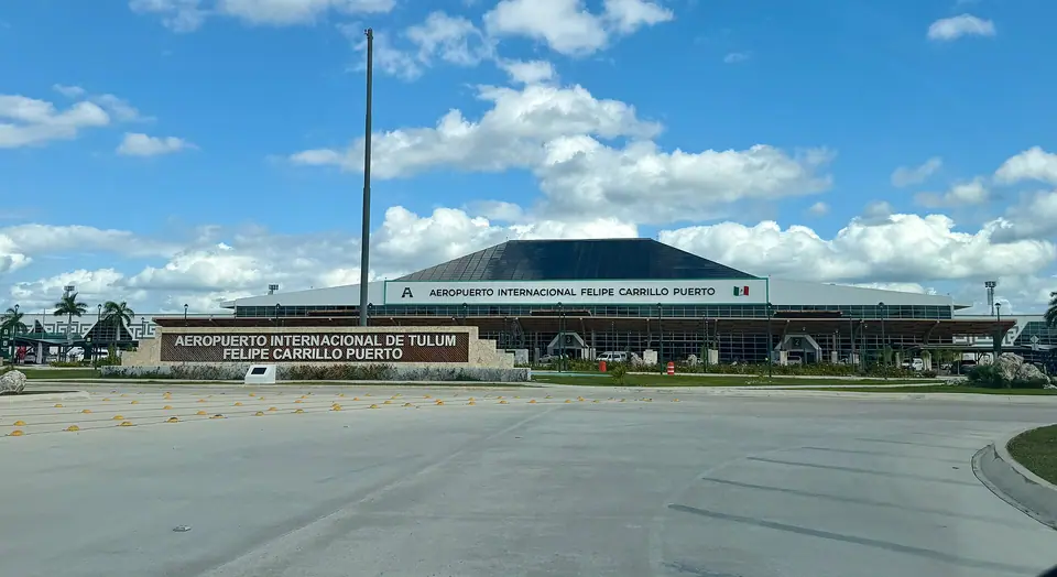 Low terminal building of Tulum International Airport with its name sign, a flagpole and an empty forecourt under a blue sky