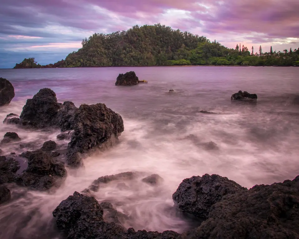 Waves on dark rocks at Hana Bay with green mountains behind