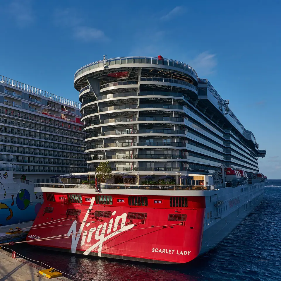 The red-hulled cruise ship Scarlet Lady docked alongside another large cruise ship at a pier in Cozumel under a blue sky