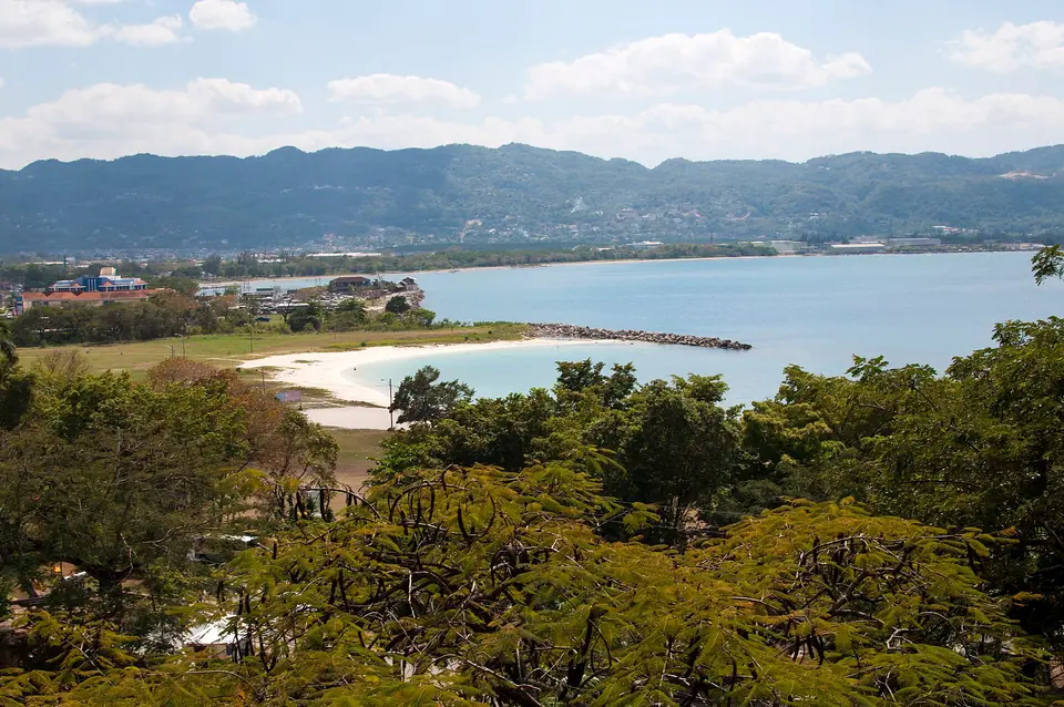 View over trees to a curving white-sand beach and breakwater on Montego Bay with green hills behind the city