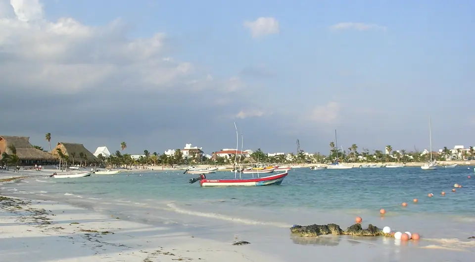 Calm, shallow Akumal Bay with a small fishing boat at anchor and low buildings and palms along the shore