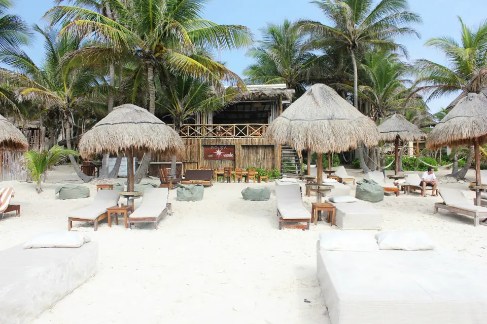 White daybeds and thatched palapa umbrellas on the sand at a beach club in Tulum's Hotel Zone, with palm trees behind