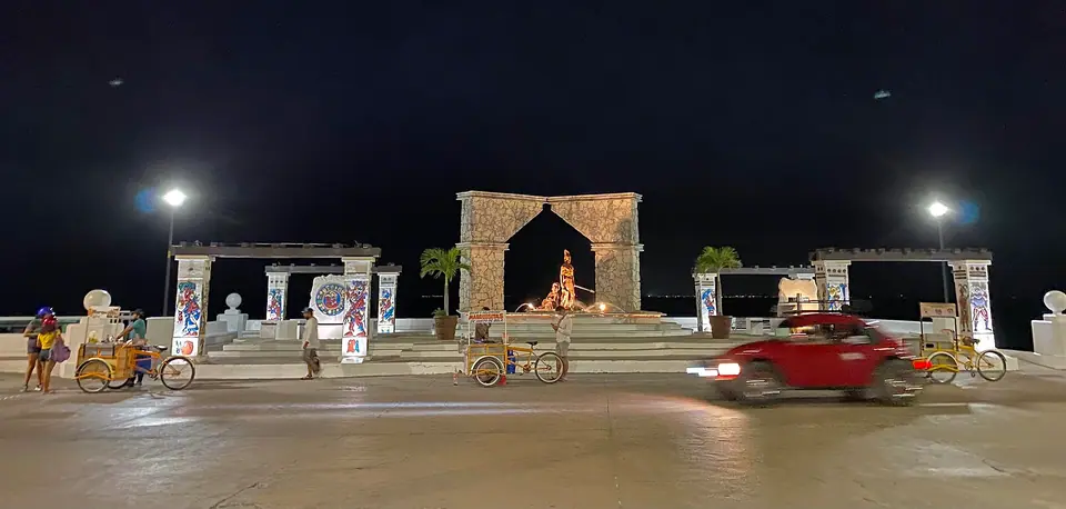 San Miguel de Cozumel's waterfront malecón at night, with a lit stone arch, streetlights and passing cars