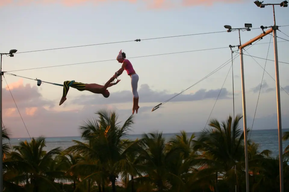 Guest swinging on the flying trapeze rig at Club Med Turkoise at dusk, with palm trees below