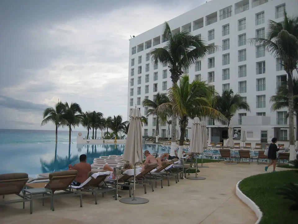 Infinity pool with loungers and umbrellas beside palm trees and the white main building of Le Blanc Spa Resort Cancun