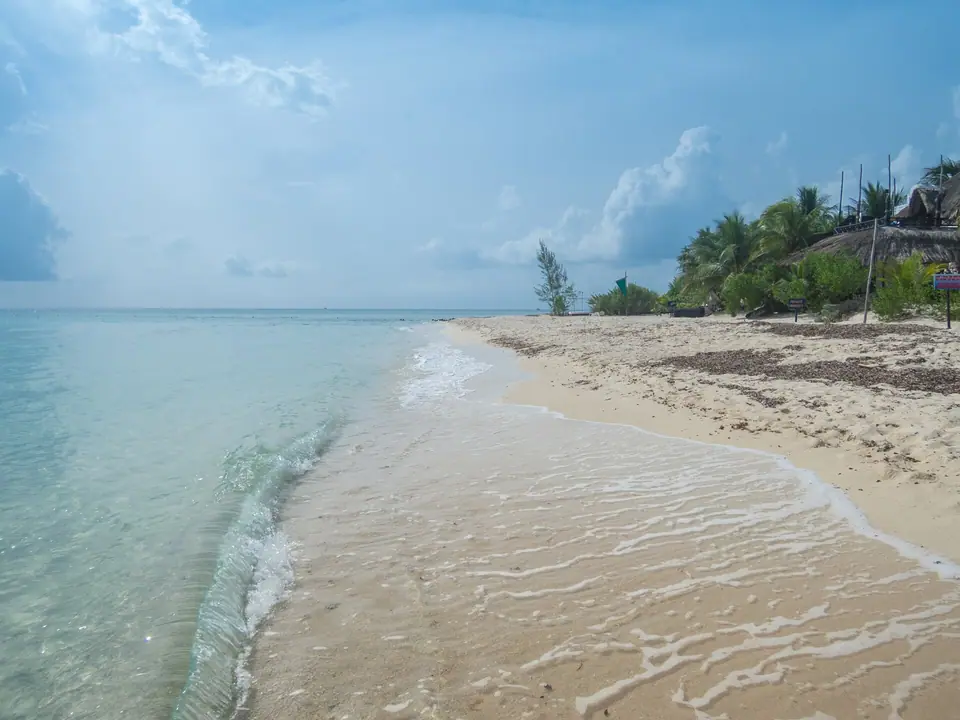 Calm, clear shallow water lapping onto pale sand at Palancar Beach on Cozumel's southwest coast, with palms and a thatched roof behind