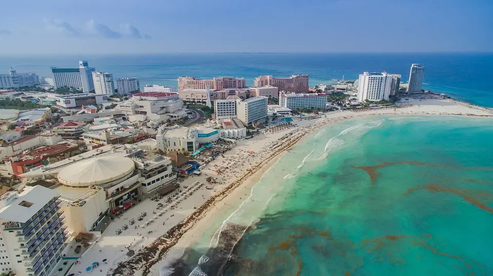 Aerial view of Cancun's Hotel Zone with high-rise resorts along a white-sand beach and turquoise sea marked by brown seaweed