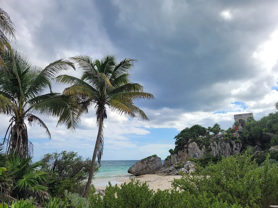 Palm trees framing the Tulum ruins on a rocky headland above the sea under a cloudy sky