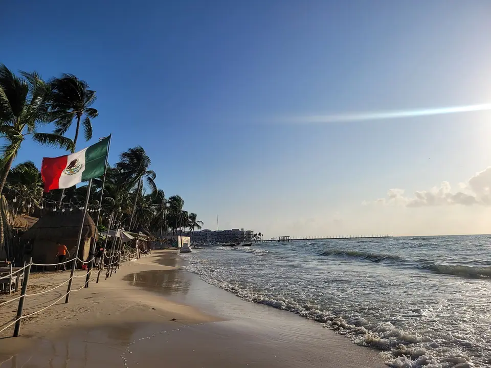 Palm-lined beach in Playa del Carmen with a Mexican flag and gentle surf at sunset