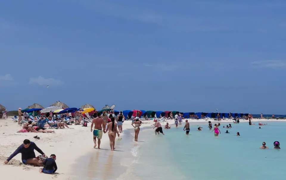 Busy public Baby Beach in southern Aruba with swimmers in shallow clear water and colorful umbrellas along the sand