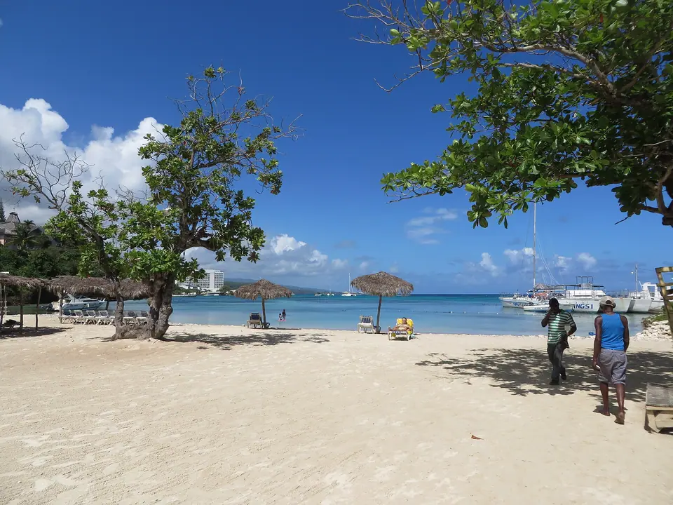Sunny Mahogany Beach in Ocho Rios, Jamaica, with shade trees, thatched umbrellas and boats offshore