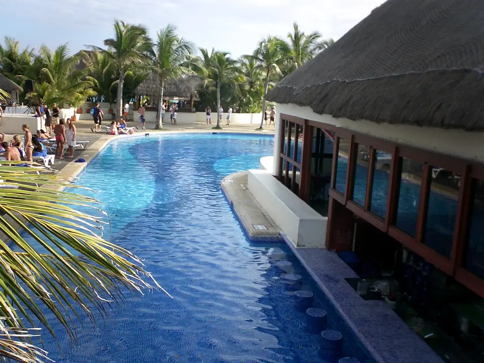 Long blue swimming pool lined with palm trees and a thatched-roof building at Grand Oasis Cancun